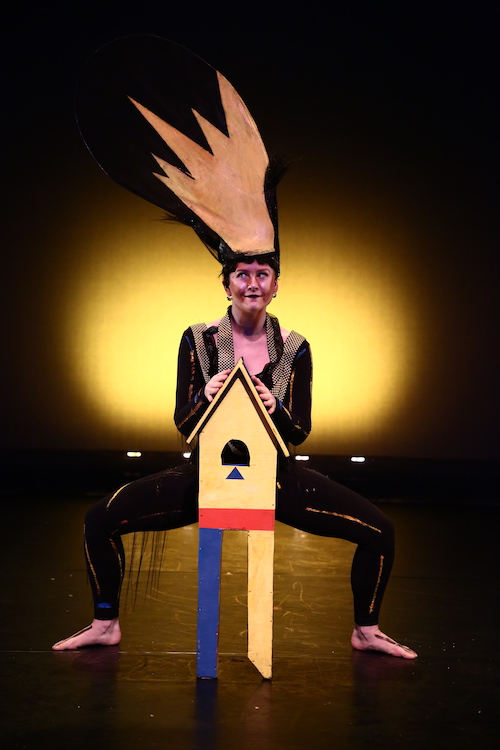 Lindsay Reuter assumes a deep second position. A small prop that looks like a birdhouse sits in front of her. She hears a headpiece with looks like painted yellow hair sticking straight up.
